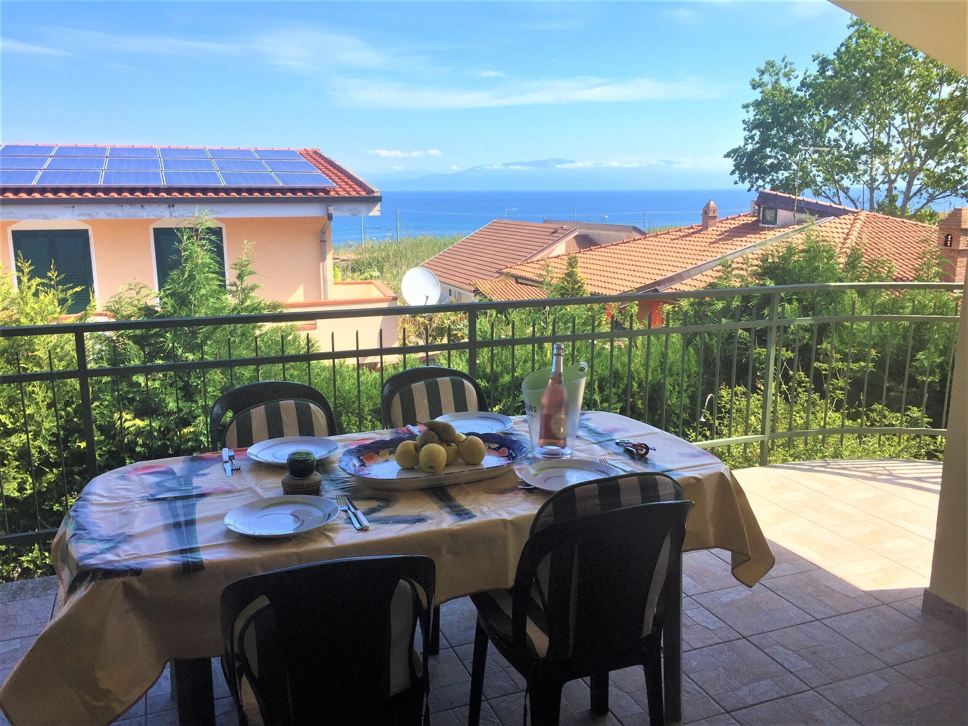 Tavolo e sedie su un balcone, con una bottiglia e piatti, vista su una zona costiera con case e cielo sereno.