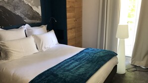 Una cama bien hecha con sábanas blancas y una manta azul, un cabecero de madera, un cuadro de montañas en la pared y una lámpara junto a la ventana.