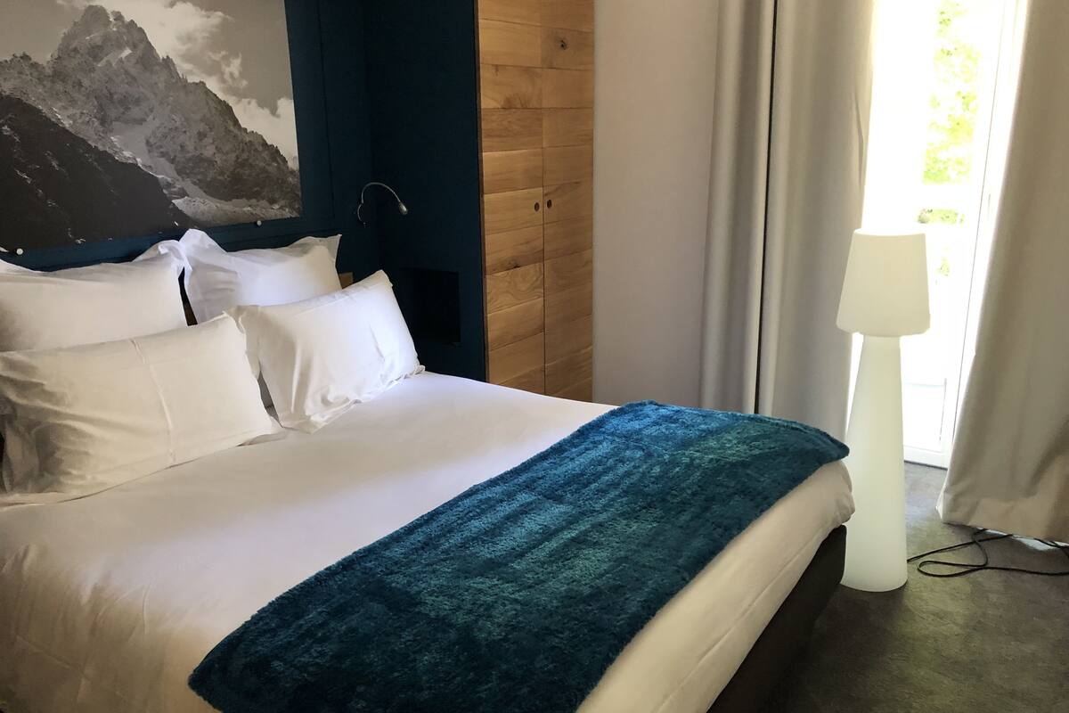 A neatly made bed with white linens and a blue blanket, a wooden headboard, a mountain painting on the wall, and a lamp by the window.