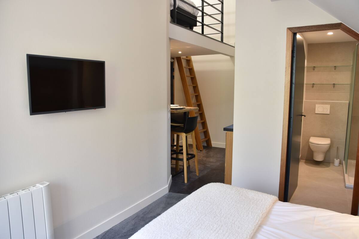A bedroom with a bed, a TV mounted on the wall, a desk with a chair, and a bathroom visible through an open door.