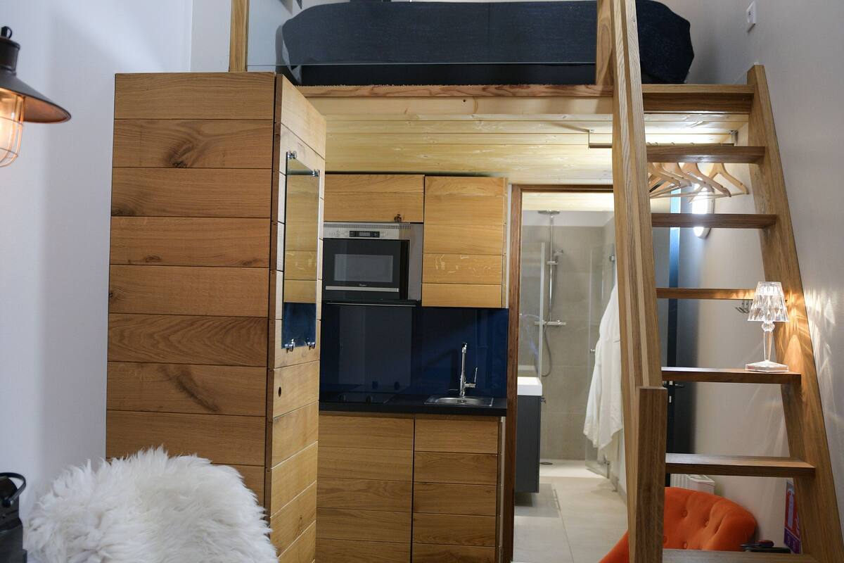 A compact room with a loft bed, a small kitchenette, a wooden wardrobe, and a seating area with a fluffy white rug and an orange chair.