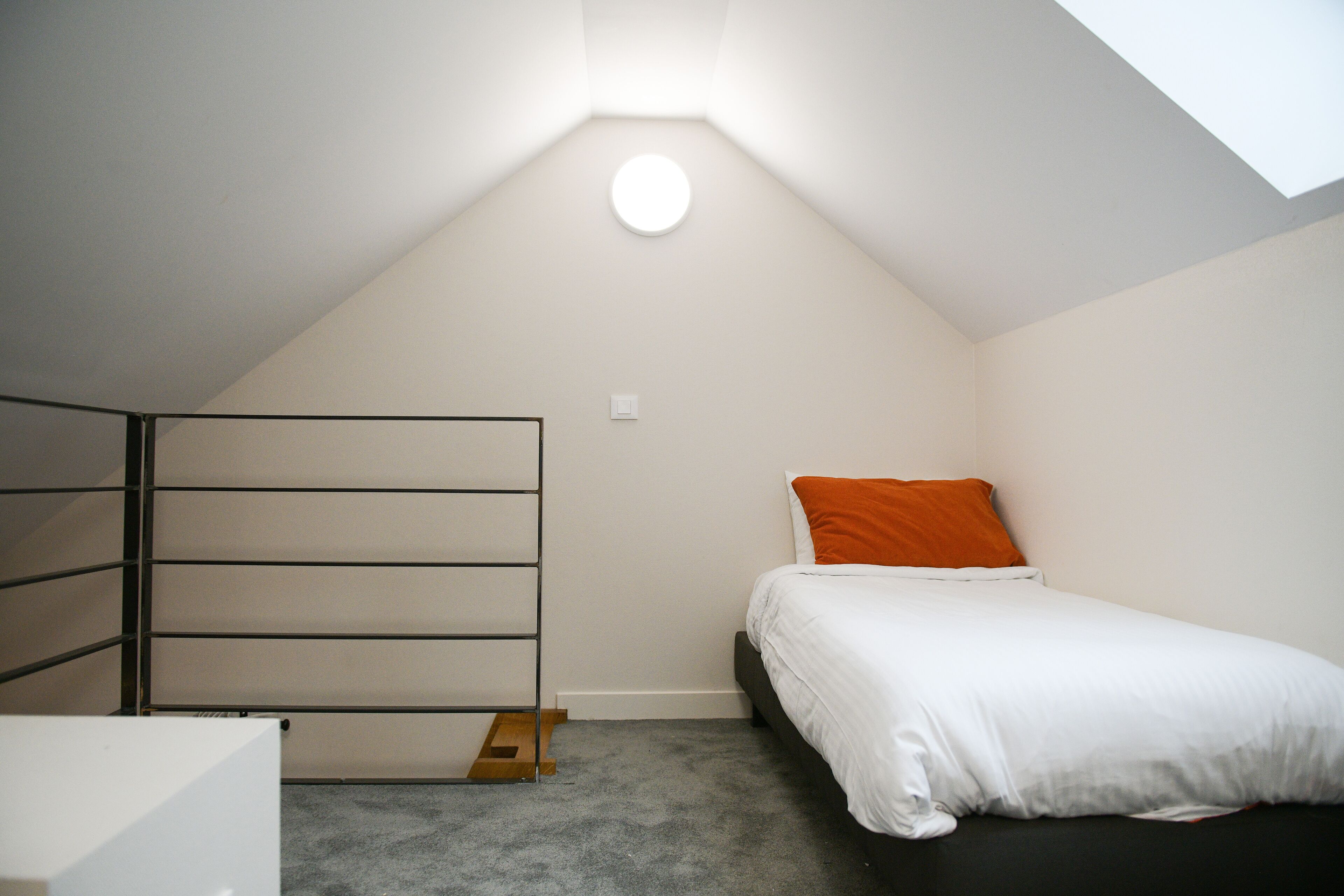 A small, well-lit room with a sloped ceiling, a bed with white bedding and an orange pillow, and a metal rack against the wall.