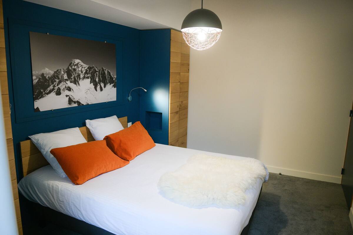 A bedroom with a bed, white and orange pillows, a white fluffy blanket, a mountain photograph on the wall, and a modern pendant light.