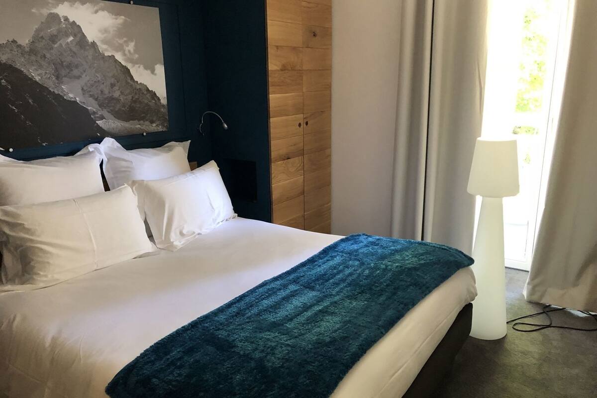 A neatly made bed with white linens, a blue throw blanket, and a mountain painting on the wall.