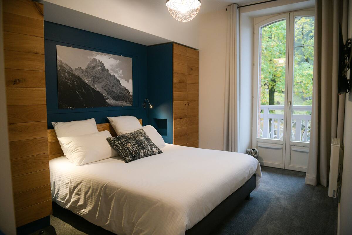 A bedroom with a bed, a large framed picture, a nightstand, and a window with a view of greenery.