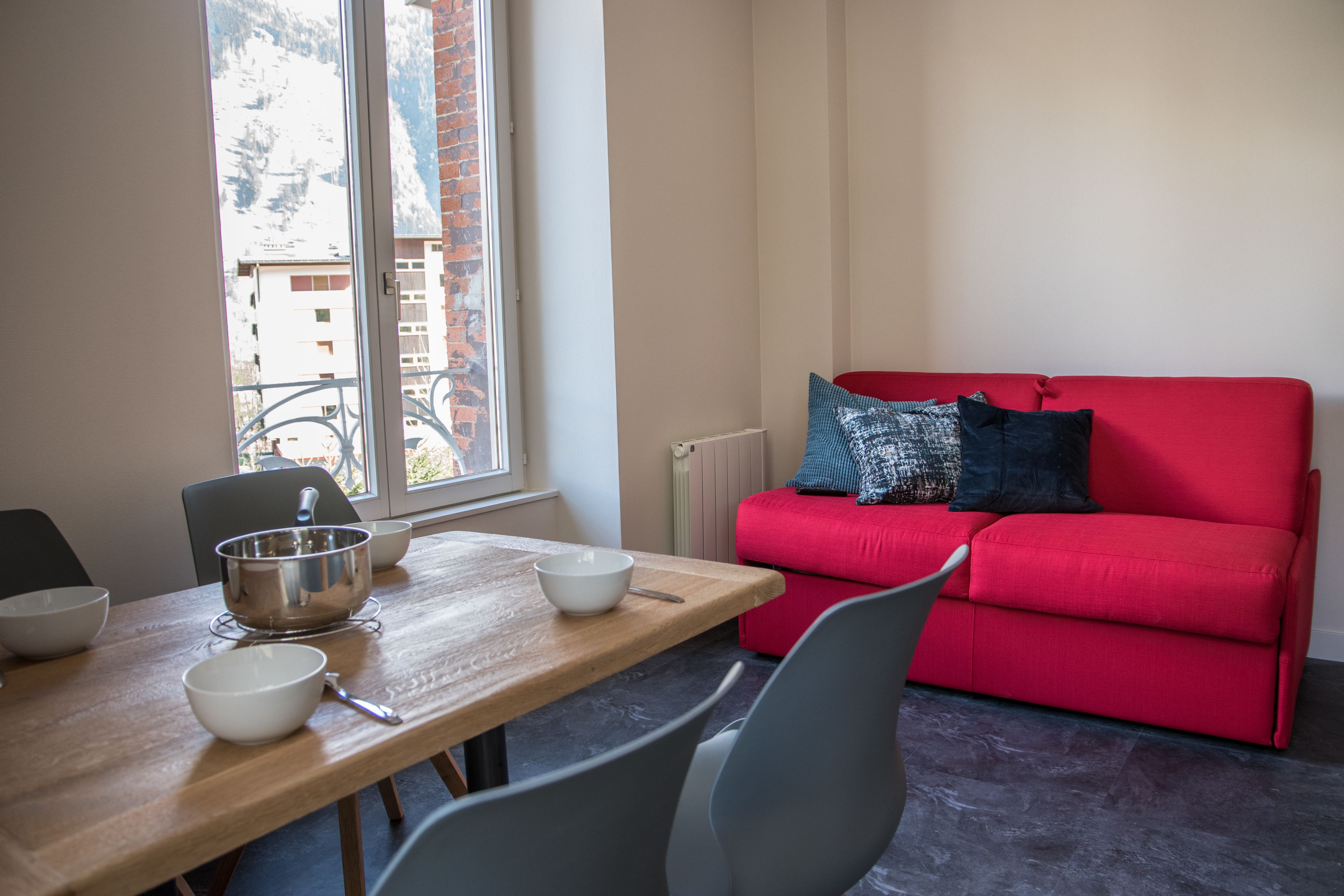 Un comedor con una mesa de madera, sillas y cuencos. Un sofá rojo con cojines estampados está apoyado en la pared.