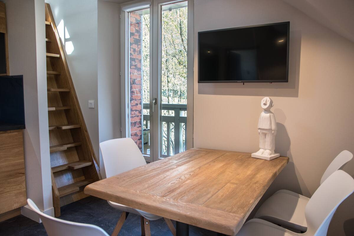 A dining area with a wooden table, white chairs, a TV mounted on the wall, and a staircase leading to an outdoor area.
