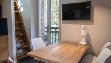 A dining area with a wooden table, white chairs, a TV mounted on the wall, and a staircase leading to an outdoor area.