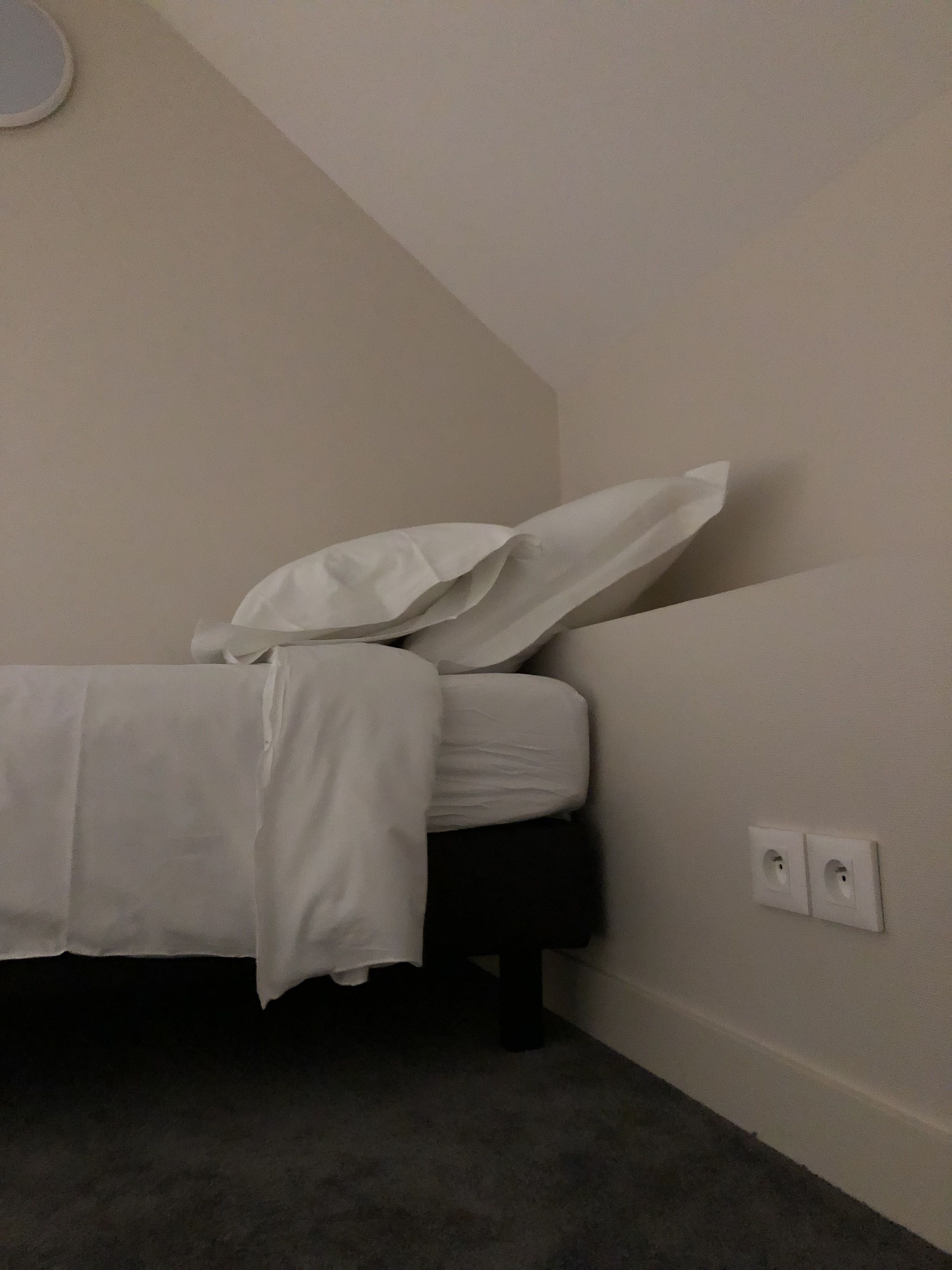 Una habitación pequeña y con poca luz, con una cama individual, un enchufe y suelo cubierto de alfombra.