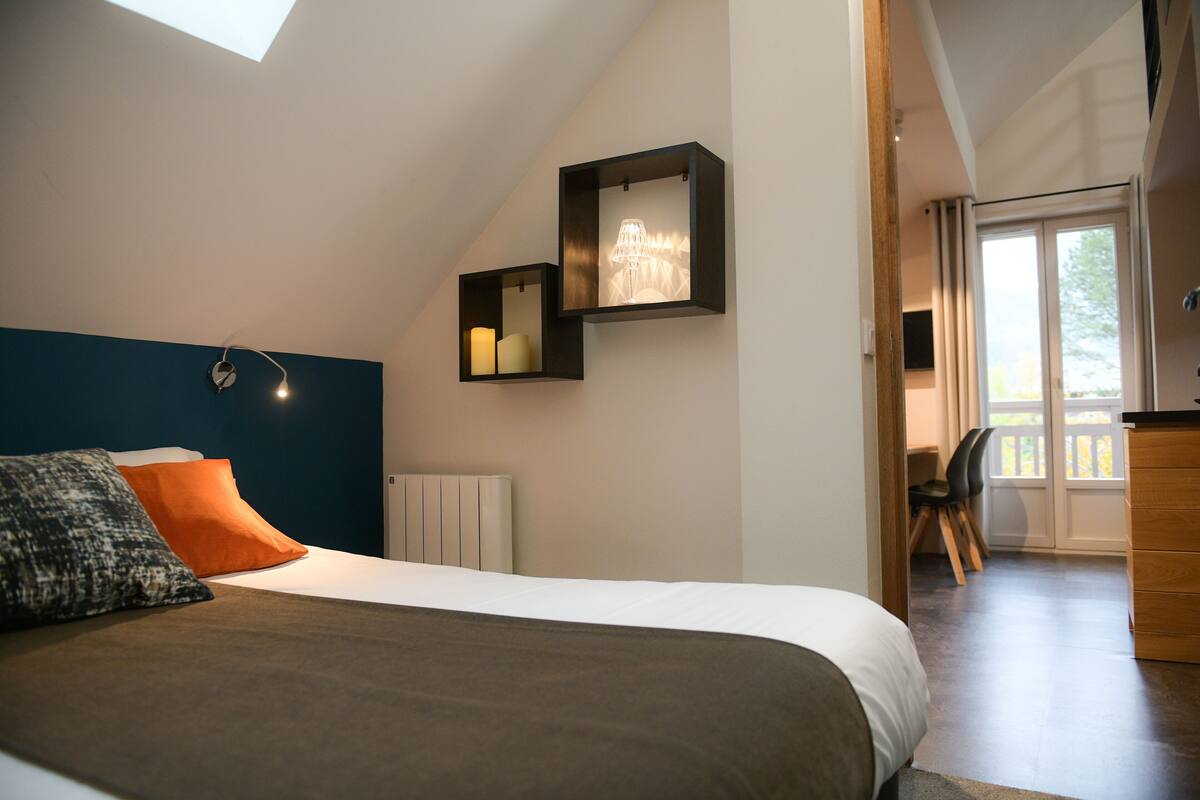 A bedroom with a sloped ceiling, a bed with a white and brown bedspread, two pillows, a nightstand with a lamp, and a view of a balcony through a door.