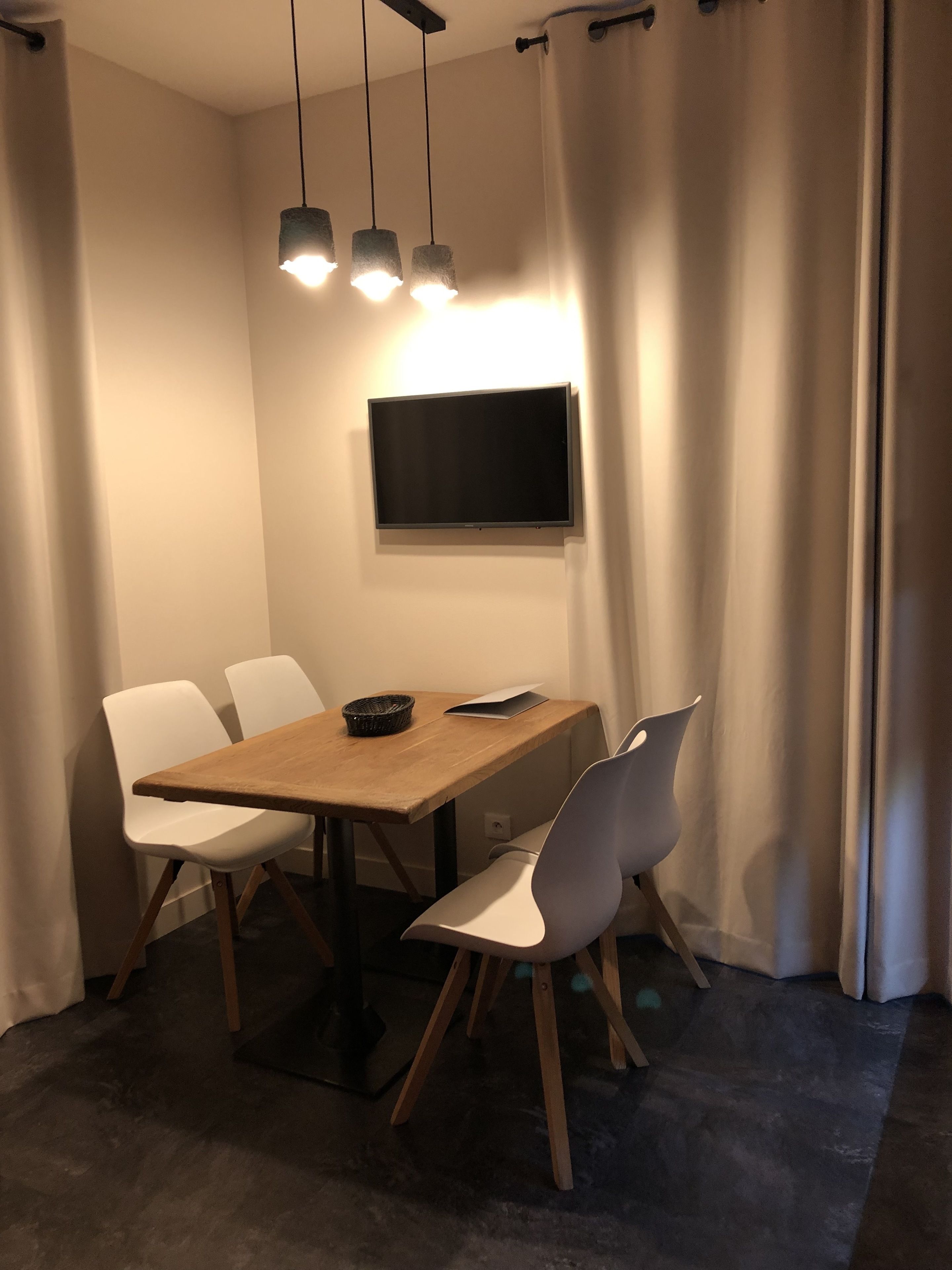 Un coin repas avec une table en bois, des chaises blanches, un téléviseur à écran plat fixé au mur et des lampes suspendues.