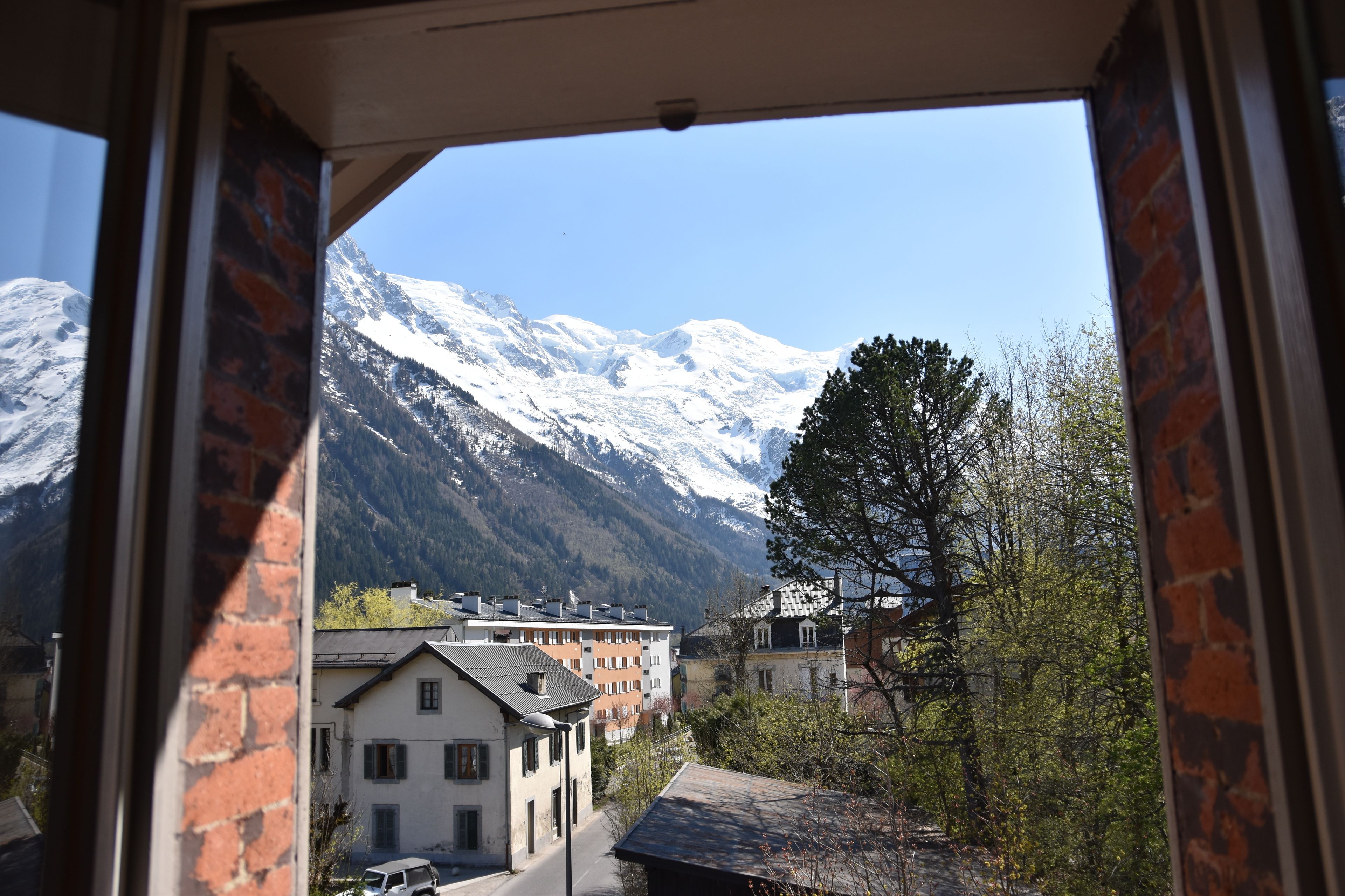 Blick durch ein Fenster auf ein Gebäude mit einem schneebedeckten Berg im Hintergrund, einem Baum und mehreren Gebäuden unterschiedlicher Architektur.
