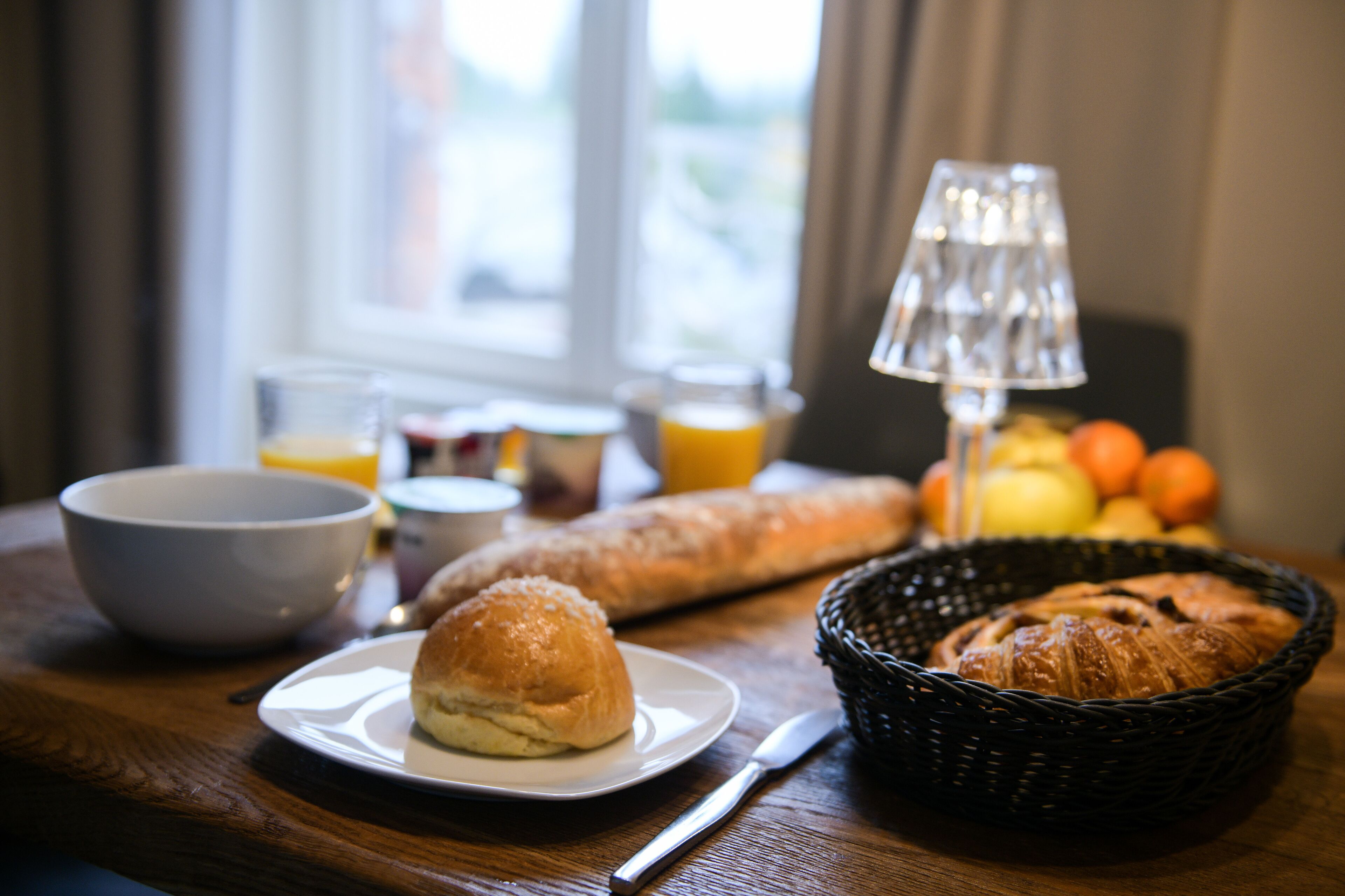 A breakfast setting with bread, a basket of fruits, a bowl, and a glass of orange juice on a wooden table.