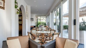 A spacious indoor area with a round table surrounded by wicker chairs, a sofa with cushions, and a large window providing a view of the outdoors. - Hotel Azzurra (Pietrasanta)