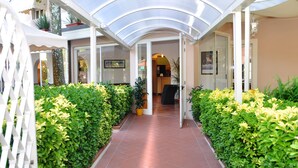A covered walkway with a glass ceiling, flanked by neatly trimmed hedges and potted plants. - Hotel Azzurra (Pietrasanta)