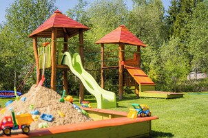 Un parque infantil con dos juegos, incluyendo toboganes y zonas de escalada, rodeado de césped y árboles.