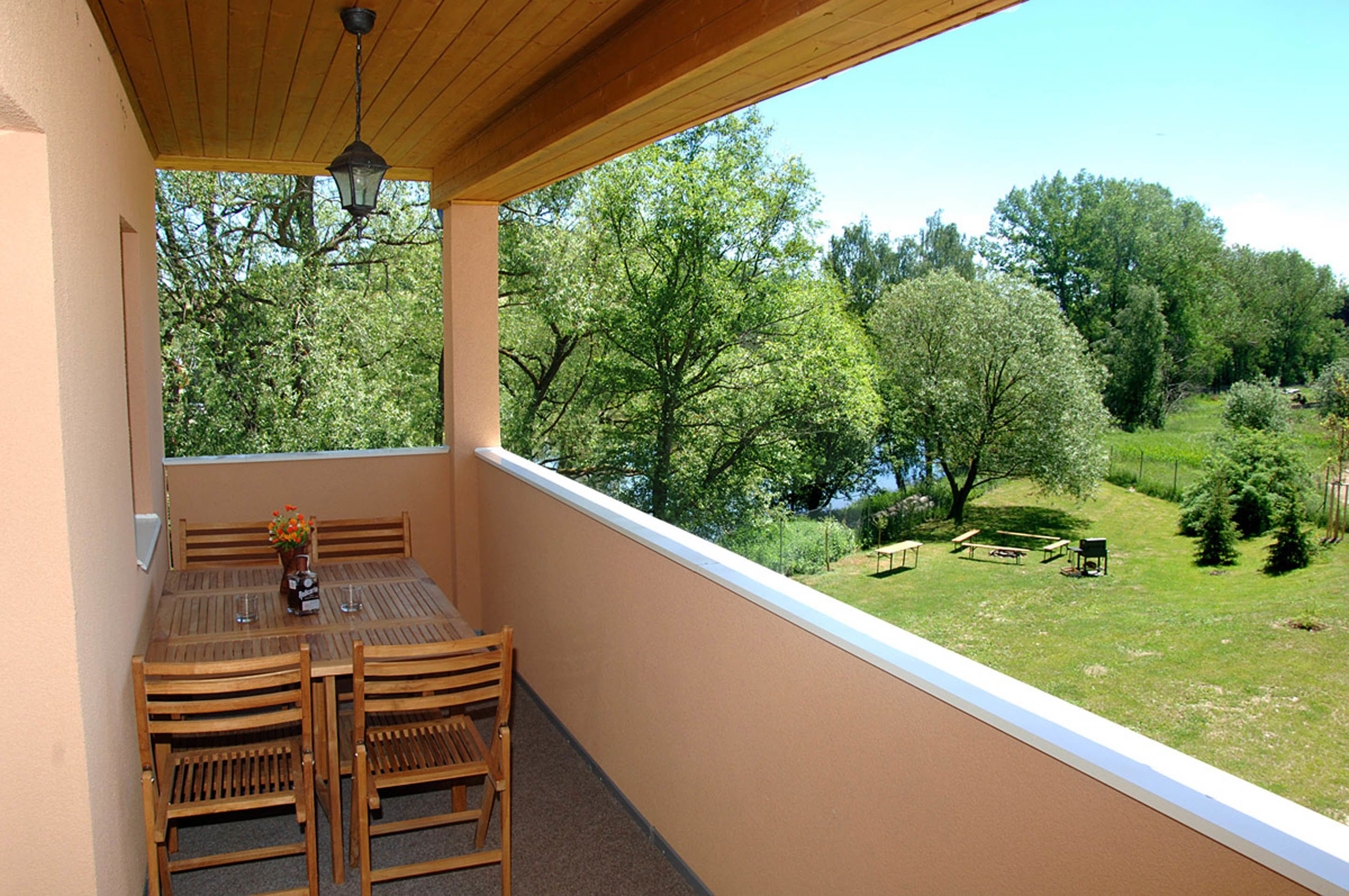 A balcony with a wooden table and chairs, a hanging light fixture, and a view of a green landscape with trees and a body of water.
