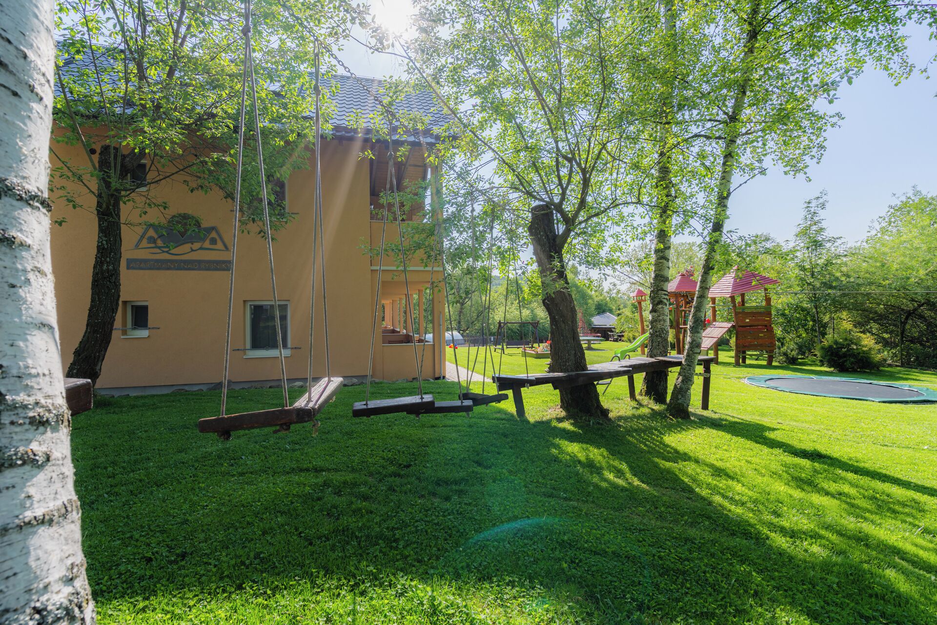 A playground with swings and a slide, surrounded by trees and a building with a yellow facade.