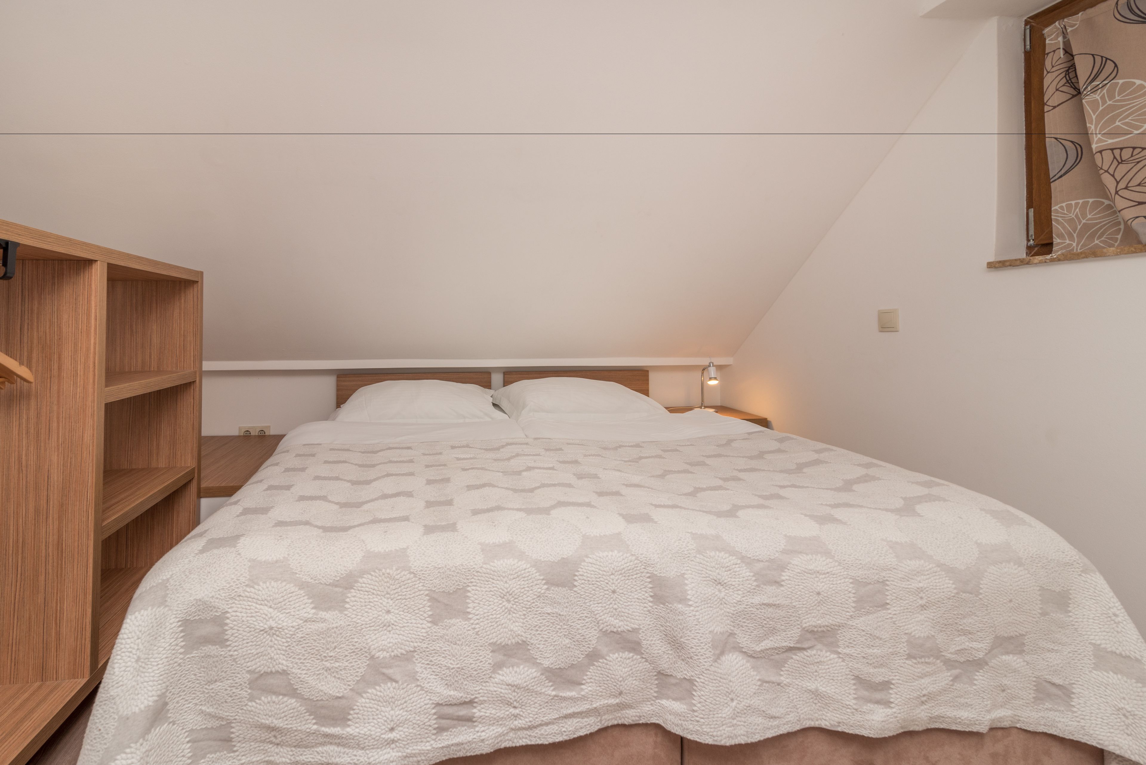 A bedroom with a sloped ceiling, a wooden wardrobe, a bed with a white floral bedspread, and a wall-mounted light fixture.