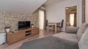 A modern living room with a stone wall, a flat-screen TV, a wooden TV stand, a grey sofa, a dining table with two chairs, and a small potted plant. - Villa Lucica Trogir (Trogir)