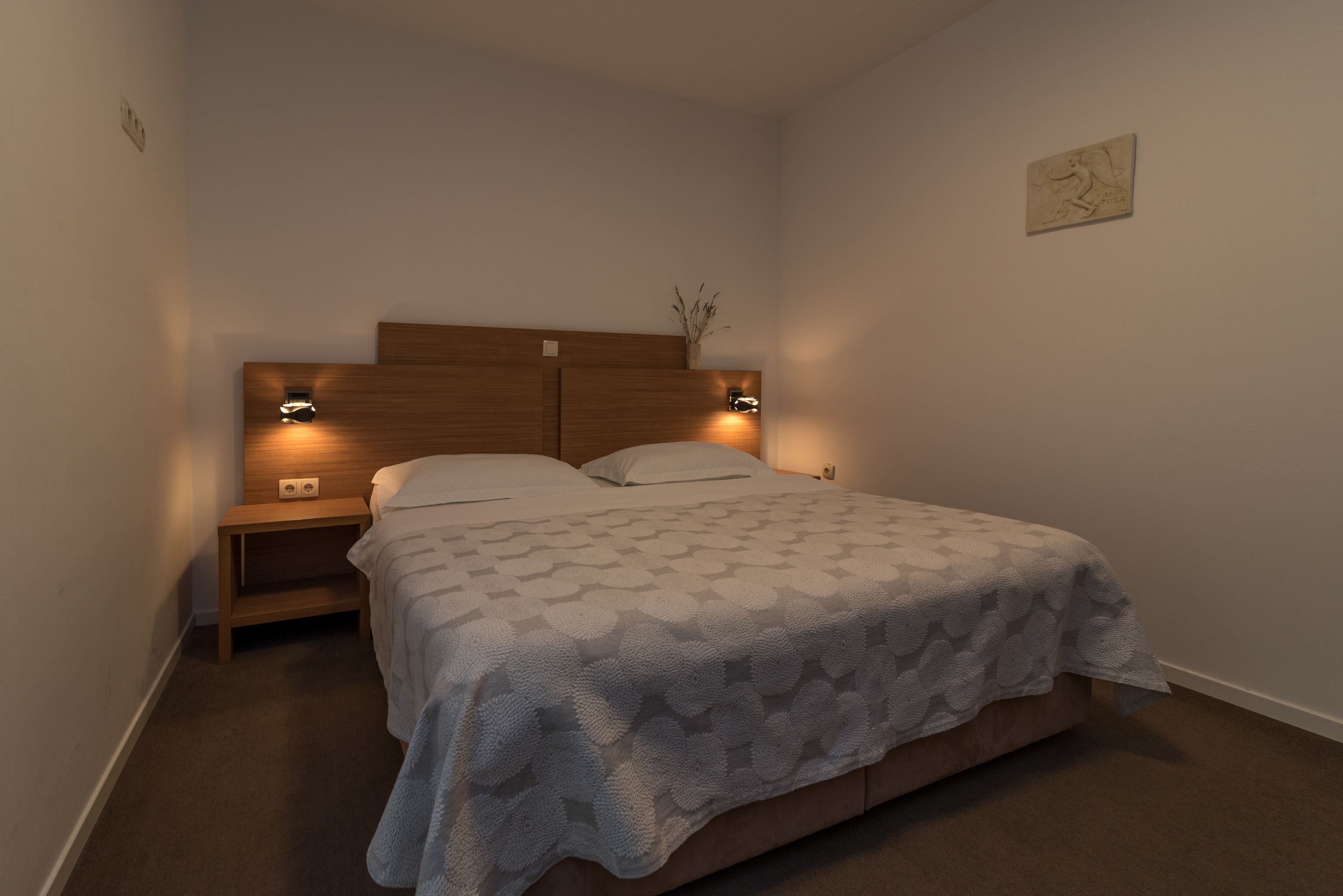 Une chambre à coucher avec une tête de lit en bois, deux tables de chevet avec des lampes, une couvre-lit à motifs et une petite œuvre d’art accrochée au mur.