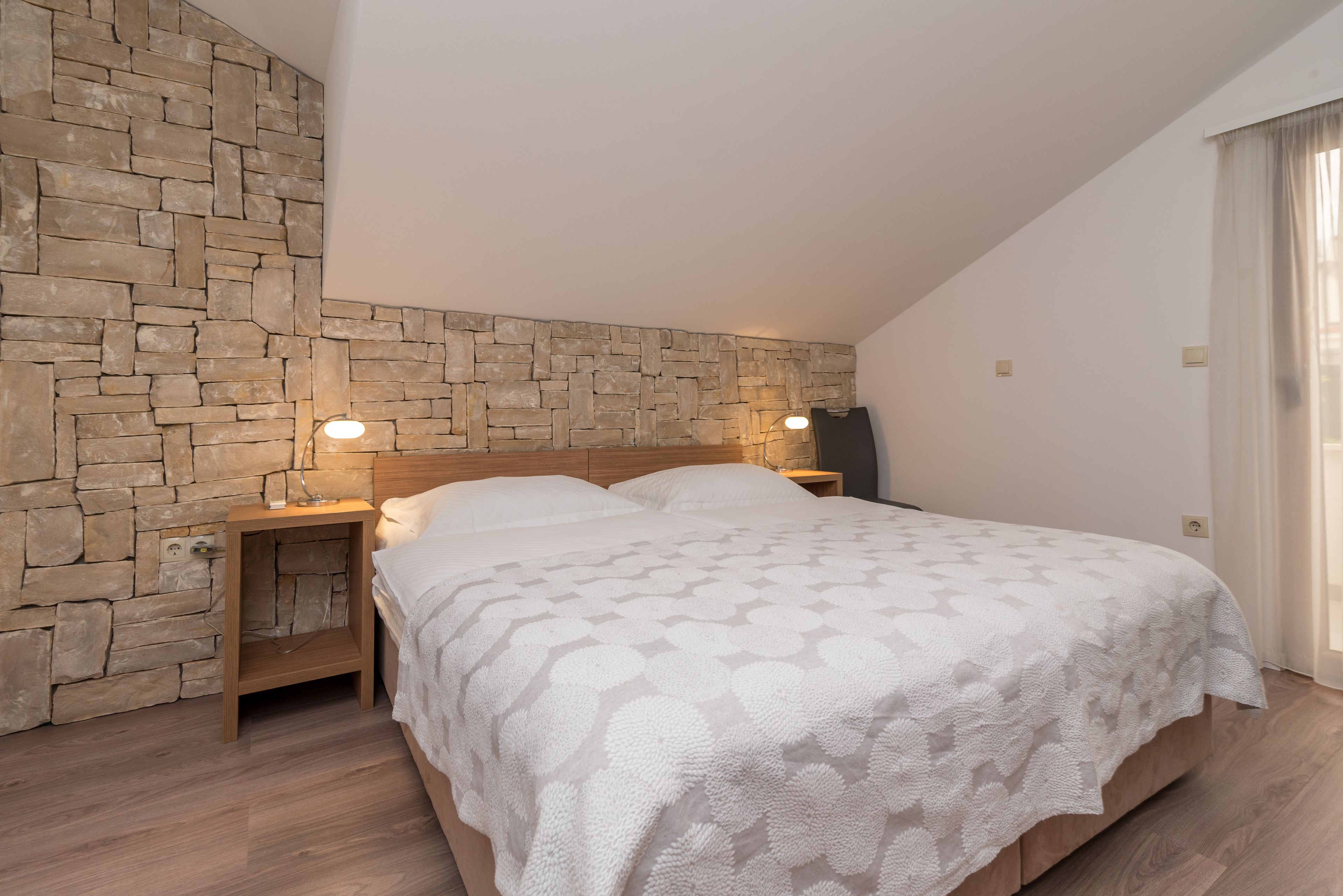 A bedroom with a stone wall, a bed with a patterned blanket, bedside lamps, and a nightstand.