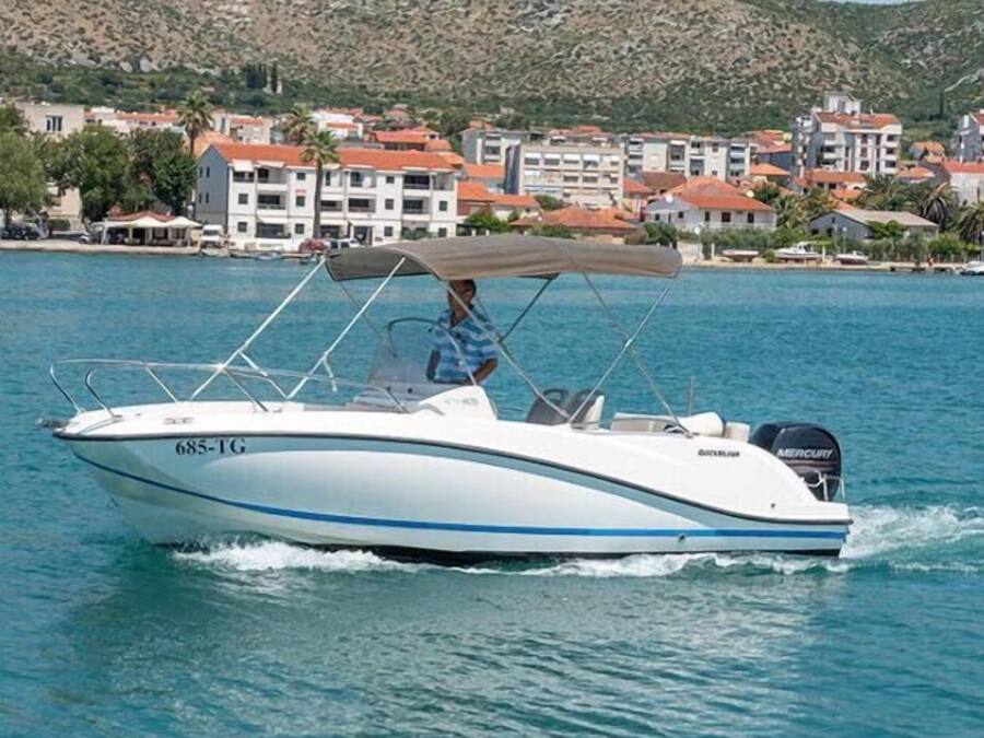 Un velero con cabina y motor navegando sobre aguas con un pueblo costero al fondo.