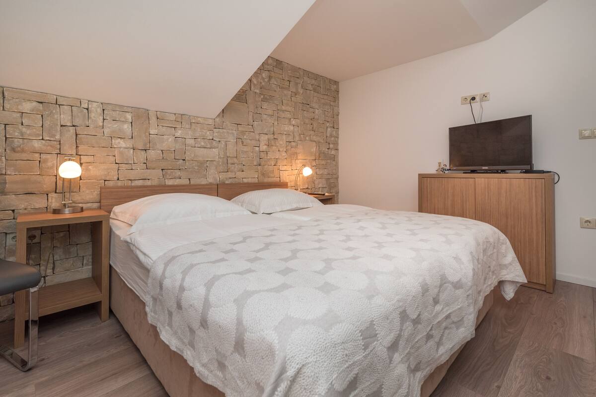 A bedroom with a stone wall, a bed with a patterned blanket, a nightstand with a lamp, and a television on a wooden cabinet.