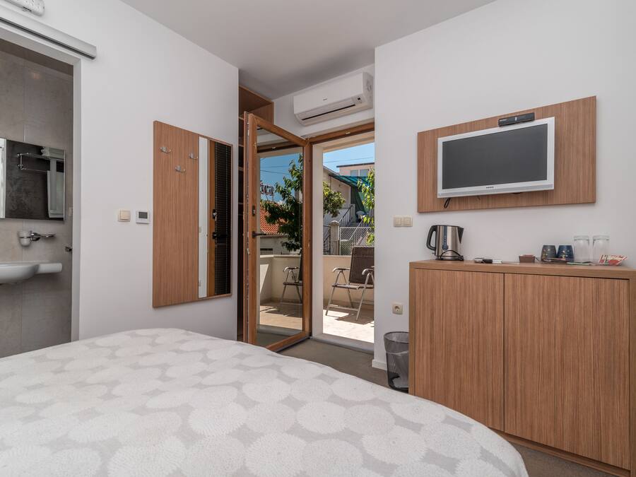 A modern hotel room with a bed, a wooden wardrobe, a television mounted on the wall, a sink, and an open door leading to a balcony.