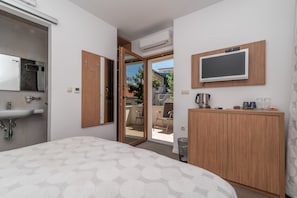 Ein modernes Hotelzimmer mit Bett, einem Holzschrank, einem an der Wand montierten Fernseher, einem Waschbecken und einer offenen Tür zu einem Balkon.
