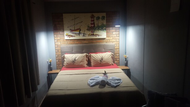 A bedroom with a brick wall, a bed with a red blanket, two pillows, a nightstand with a vase of flowers, and a painting above the bed. - Pousada Dois Corações (Penha)
