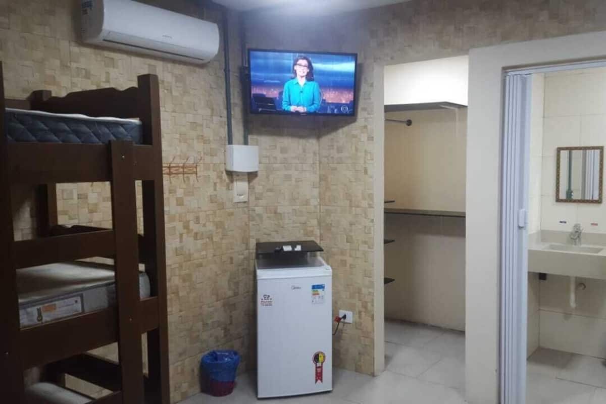 Une chambre avec un lit superposé, un petit réfrigérateur, un téléviseur fixé au mur, une salle de bain avec un lavabo et un miroir, et un sol carrelé.