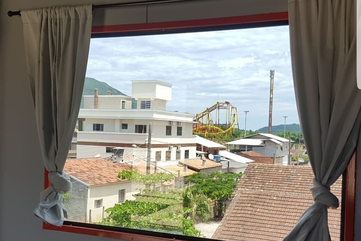 Una habitación con ventana que da a una zona residencial, con vistas a casas y una montaña rusa.