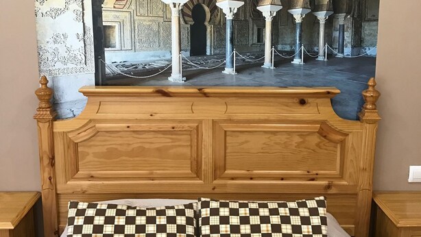 Een slaapkamer met een groot bed, een hoofdbord en een wandtapijt waarop een architectuurinterieur is afgebeeld.