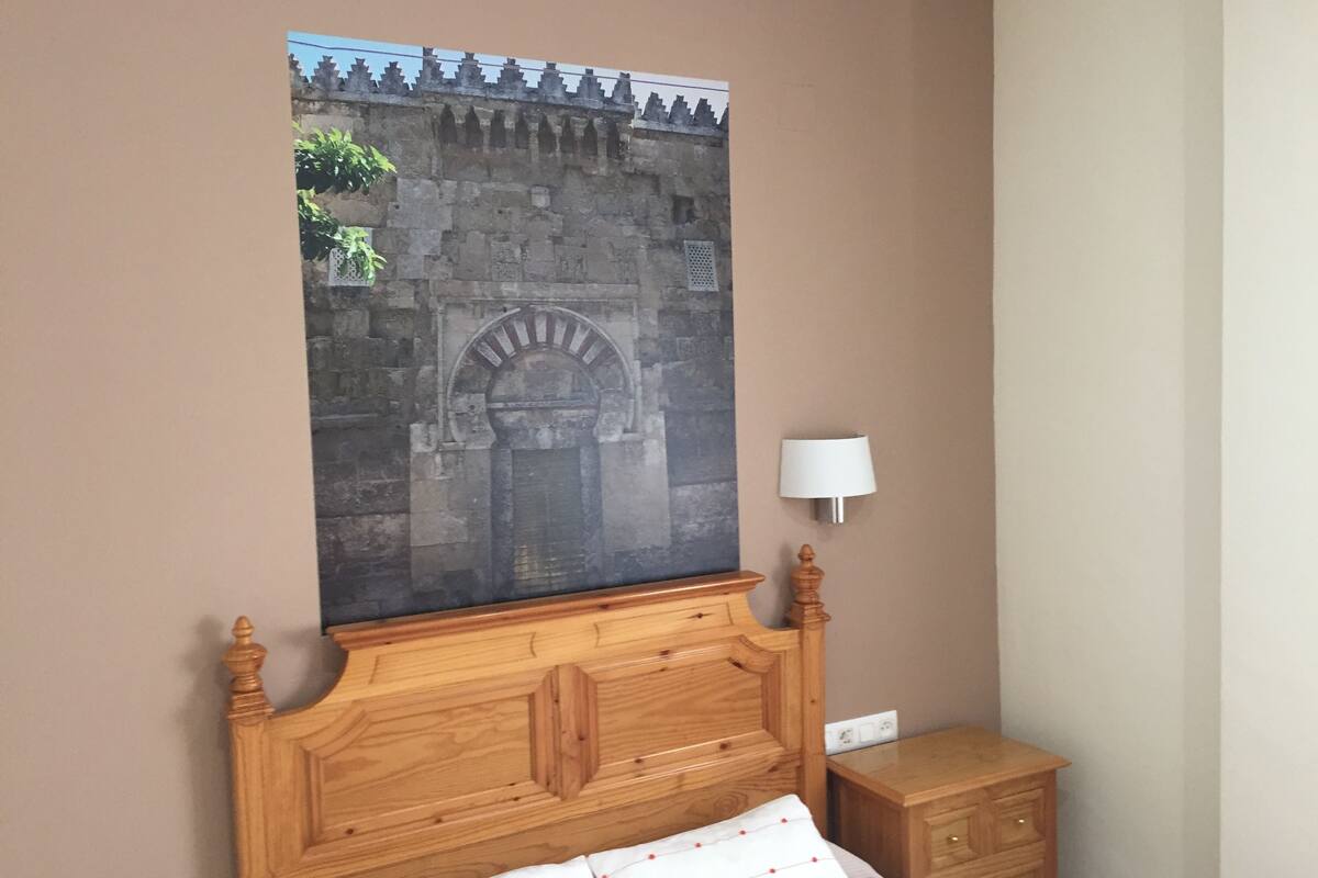 A bedroom with a wooden headboard, a bed with white bedding, a nightstand, a wall-mounted lamp, and a large poster of a stone gate.