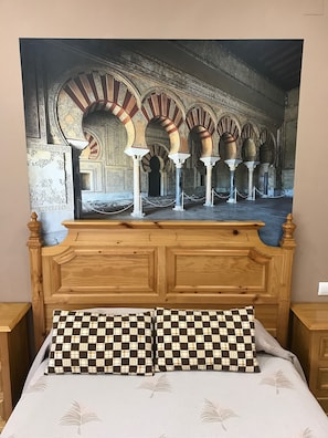 Un dormitorio con una cama grande, cabecera de madera y un tapiz que representa un interior arquitectónico.