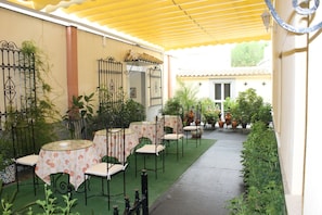 Un patio al aire libre con mesas y sillas, rodeado de plantas en macetas y un toldo amarillo.