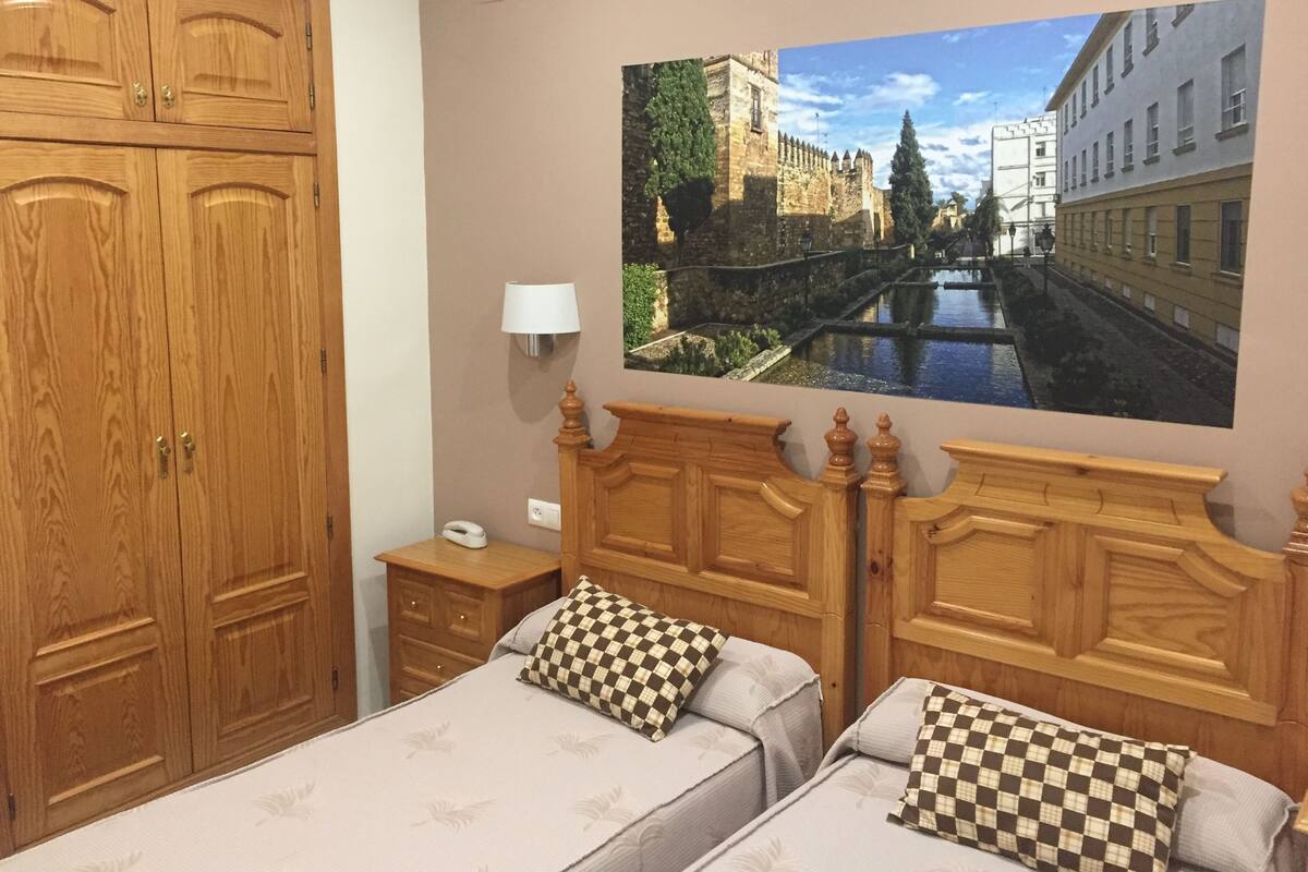 A bedroom with two beds, wooden headboards, a nightstand, a lamp, and a large wall-mounted picture of a cityscape.