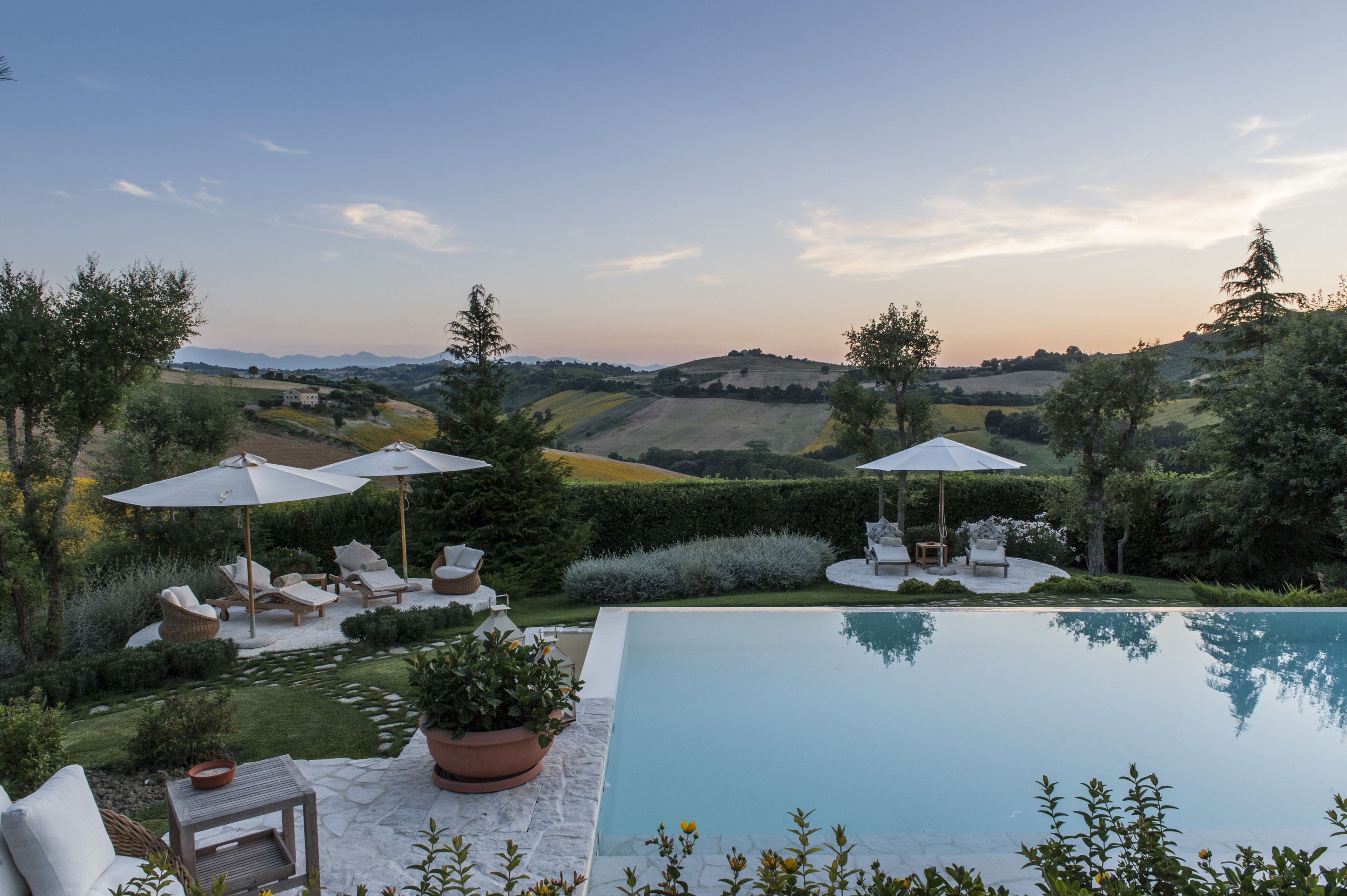 A poolside area with lounge chairs and umbrellas, surrounded by lush greenery and a view of rolling hills.