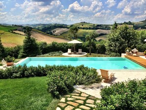 A swimming pool with a patio area, surrounded by lush greenery and hills in the background. - Monterosato Country House (Fermo)