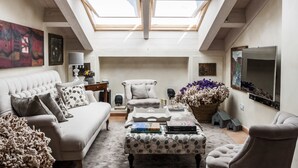 A loft-style living room with a high ceiling, skylights, a white sofa, a patterned ottoman, and a television on the wall.
