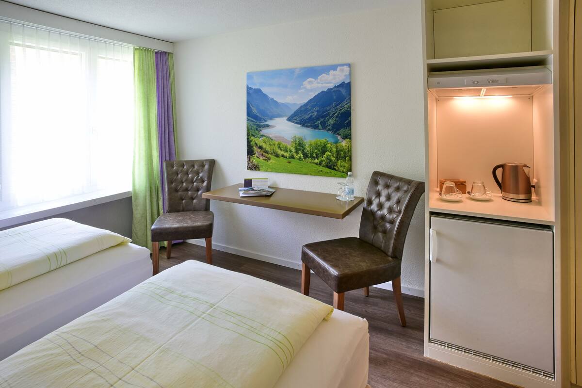 A hotel room with two beds, a dining table, two chairs, a refrigerator, and a painting of a lake.