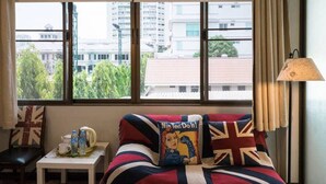 A room with a bed covered in a Union Jack blanket, a pillow with a 'Nin' character, a small table with a tray of items, and a view of buildings through large windows. - Nego Home (Bangkok)