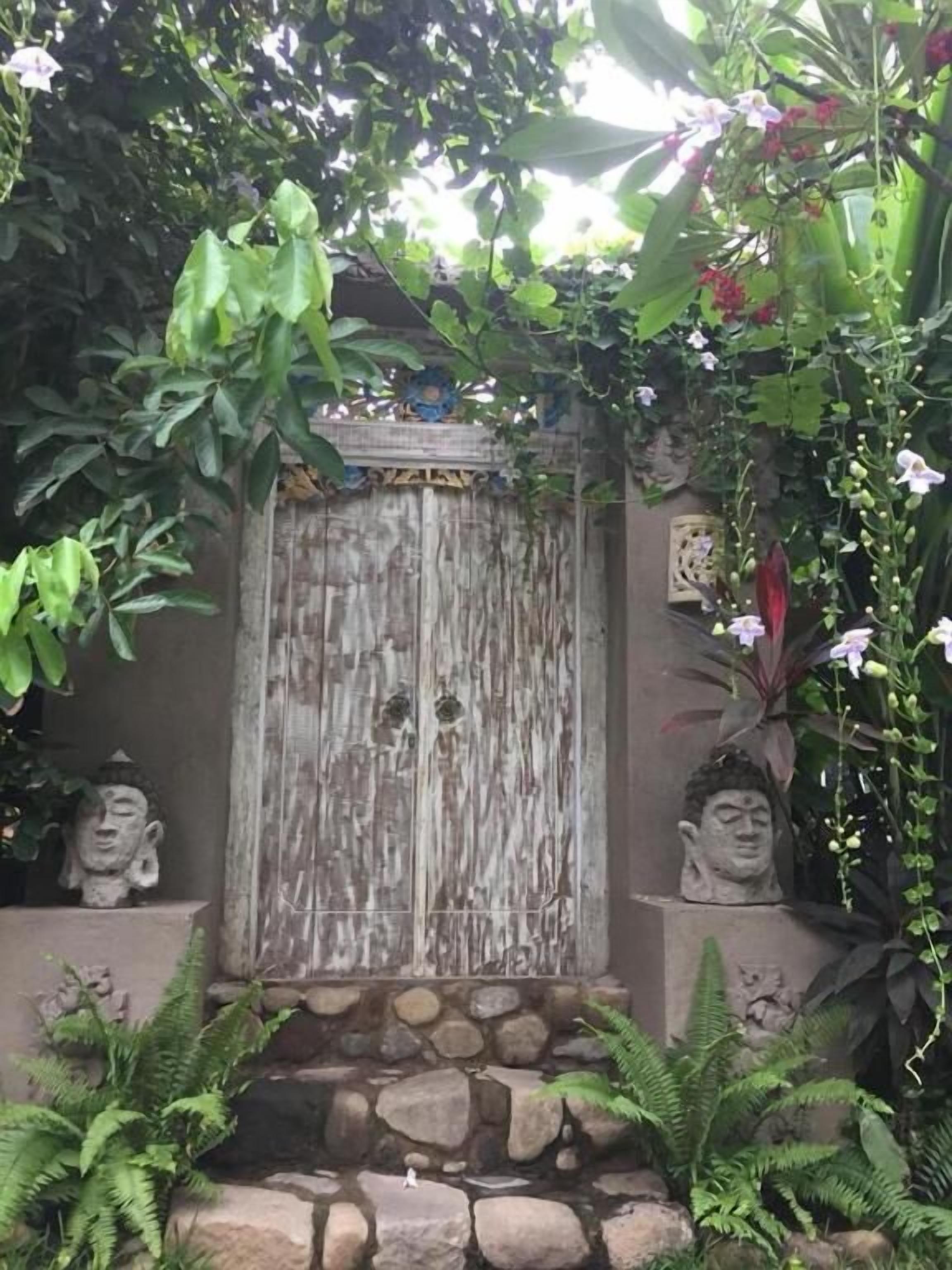 A stone wall with a wooden door, two Buddha statues, and lush greenery.