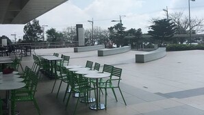 Ett utomhusområde med sittplatser, bestående av gröna stolar och bord, under en del av en byggnad som är skyddad av ett tak.