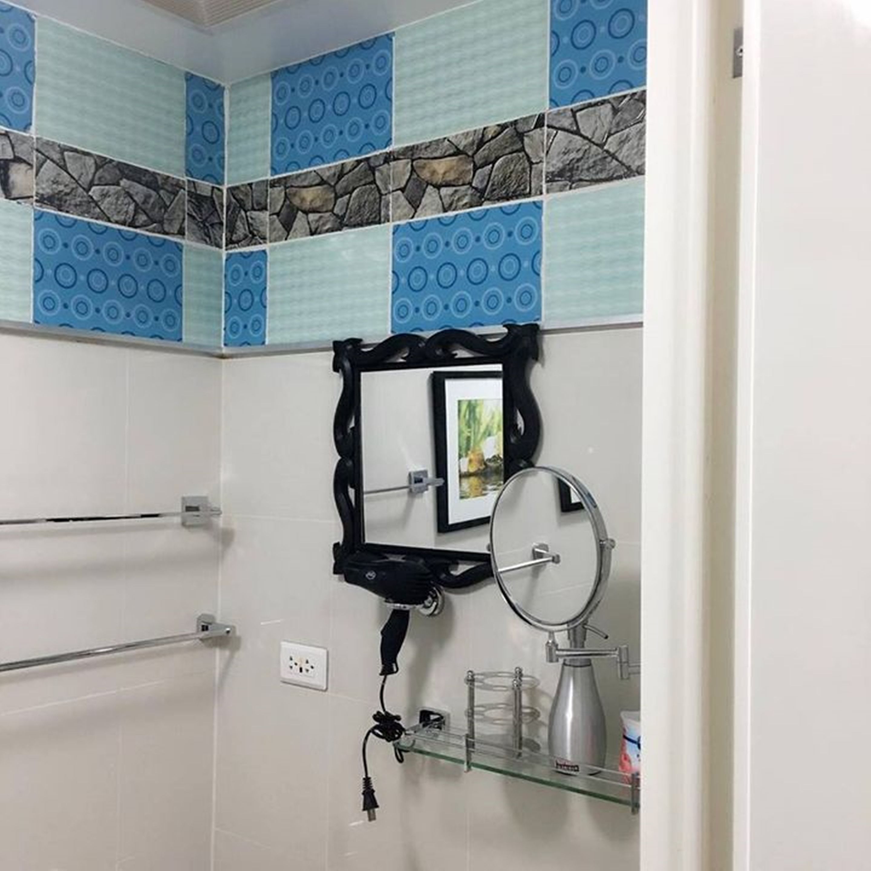 Un baño con pared de azulejos, un espejo de marco negro, un espejo redondo, un estante de vidrio con artículos de aseo y un perchero.