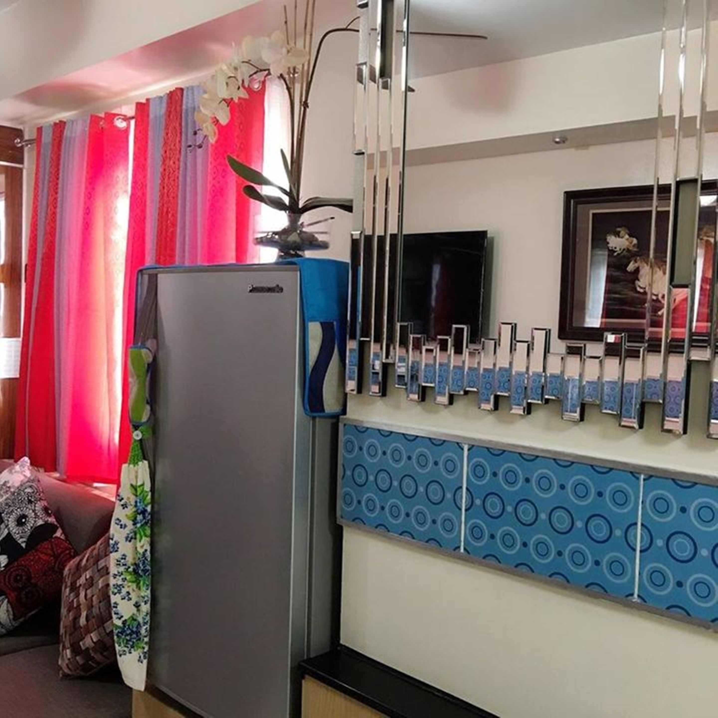 A living room with a refrigerator, a flat-screen TV mounted on the wall, and a patterned blue backsplash.