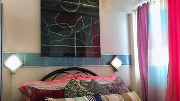 Un dormitorio con cama, almohadas, una obra de arte en la pared y una ventana con cortinas.