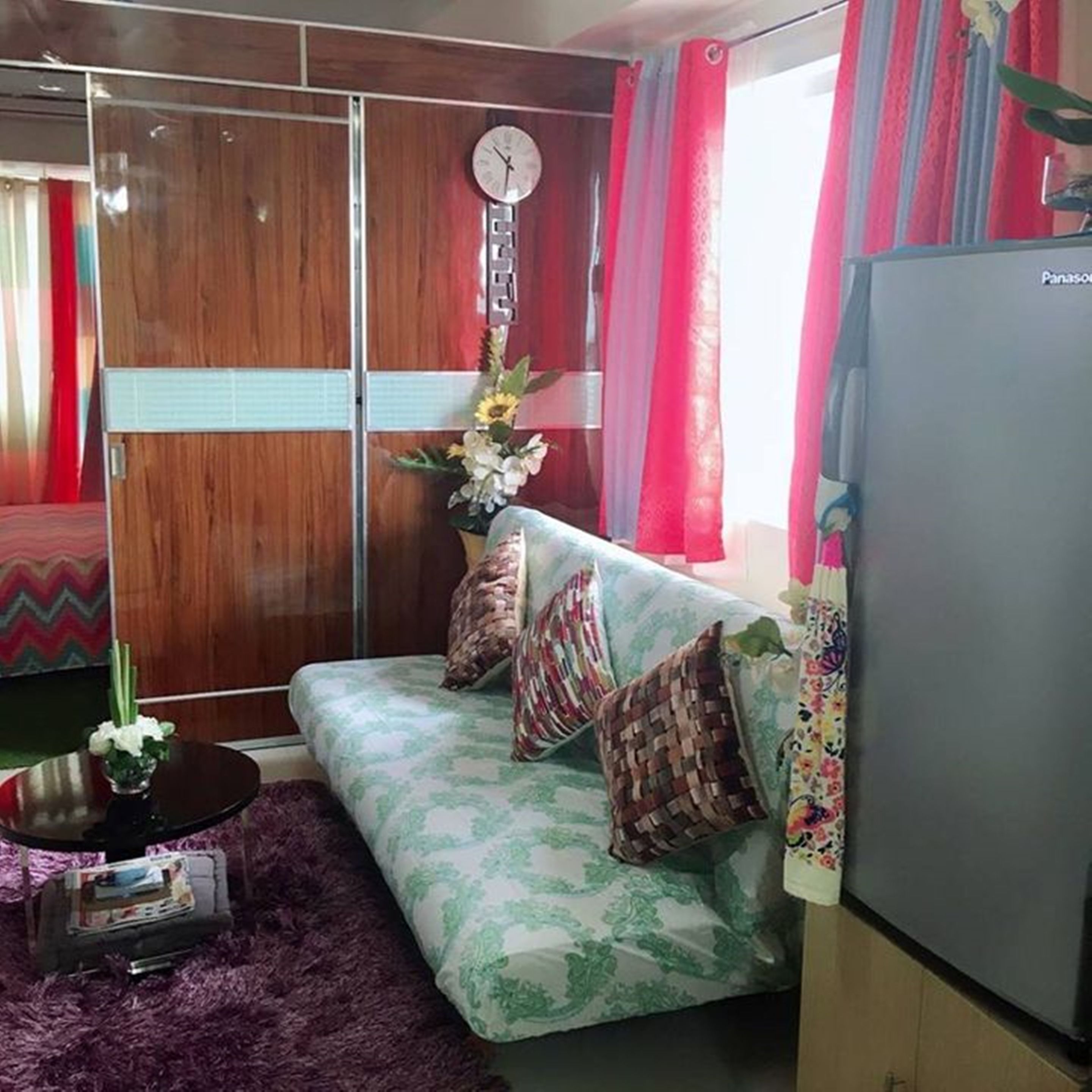 A living room with a sofa, a clock, a refrigerator, and a window with curtains.