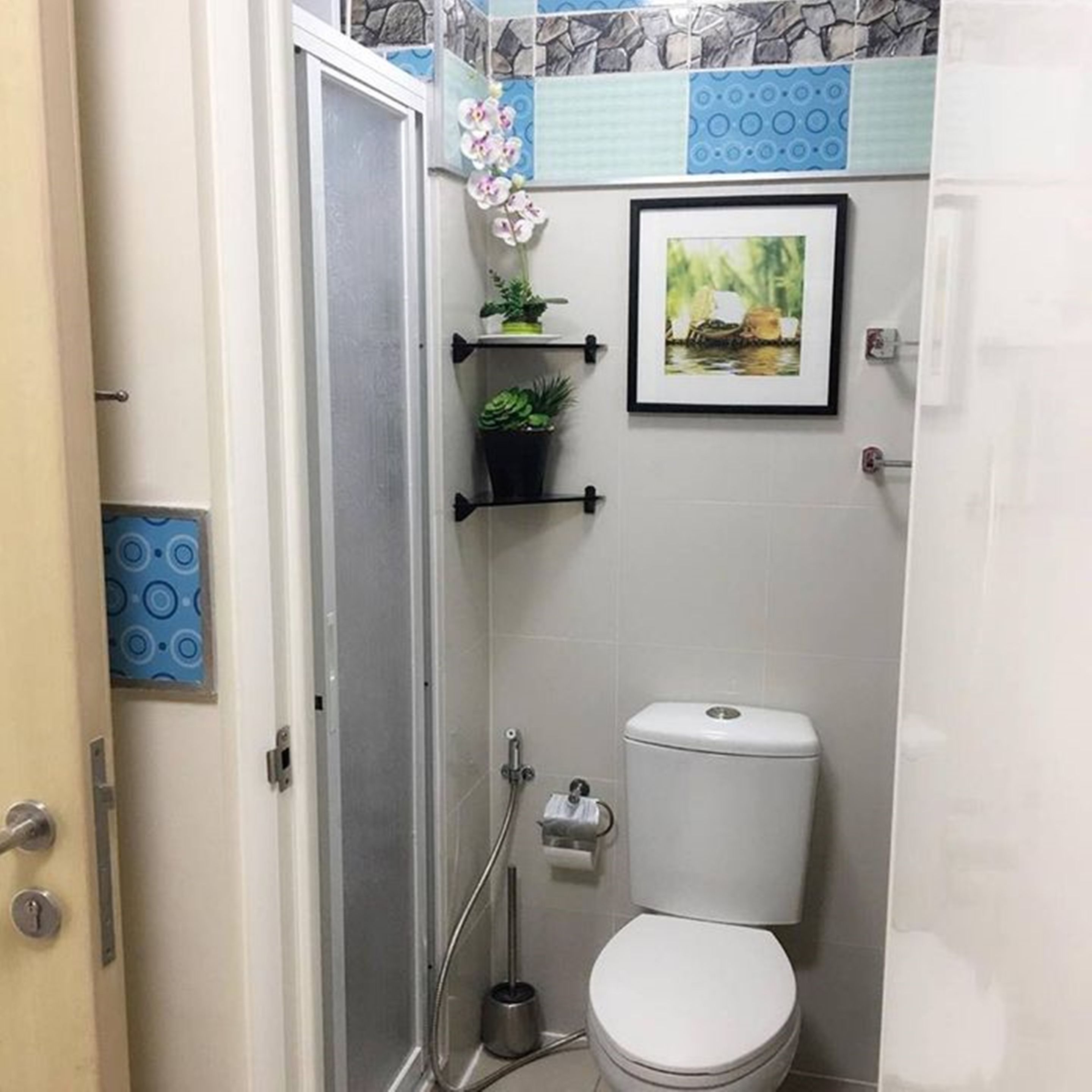 Une petite salle de bain avec des toilettes, une douche et un lavabo. Les murs sont décorés de carreaux et un tableau encadré est accroché au-dessus des toilettes.
